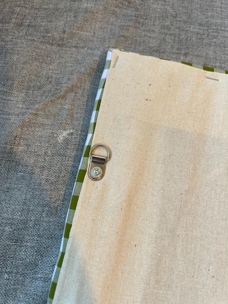 May include: Close-up of the back of a canvas art piece. The canvas is a natural beige color, with a green and white gingham pattern along the edges. A metal D-ring is attached for hanging.