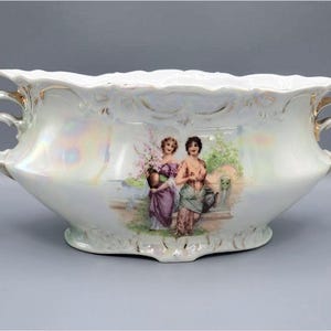 Peut inclure: Un pot en porcelaine blanche avec deux anses et un motif floral représentant deux femmes dans un jardin. Le pot a un glaçage nacré et des accents dorés.