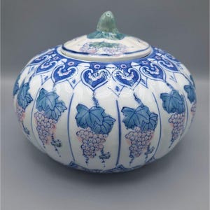 Könnte beinhalten: Eine runde Keramikdose mit Deckel, weißem Grund und blauen und rosa Traubenmustern. Die Dose hat einen blau-weiß gemusterten Rand und einen grünen Knopf auf dem Deckel. Die Dose dient wahrscheinlich dekorativen oder Aufbewahrungszwecken.