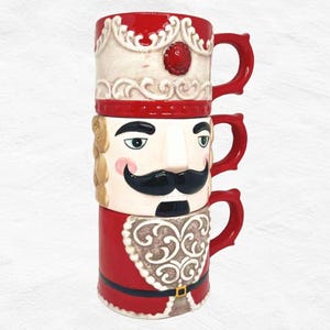 May include: A stack of ceramic mugs featuring a nutcracker design. The mugs are red, white, and beige, with detailed illustrations of a nutcracker soldier. Each mug has a red handle and is decorated with intricate patterns and facial features.
