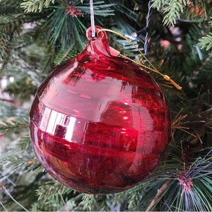 Puede incluir: Una gran bola de Navidad redonda de cristal rojo con un patrón de remolino. El adorno cuelga de un hilo dorado y está rodeado de agujas de pino verdes. El cristal refleja la luz.