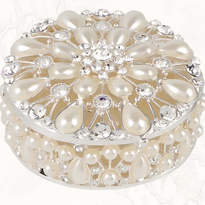 May include: A round, decorative jewelry box with a silver-colored metal frame. The box is adorned with faux pearls and clear, sparkling crystals. The lid and base are embellished with pearl and crystal accents, creating a luxurious appearance.