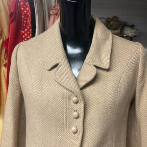 Puede incluir: Una blazer beige con un patrón de espiga, exhibida en un maniquí negro. La blazer tiene un cuello con solapas y tres botones a juego. Esta chaqueta clásica es adecuada para diversas ocasiones.