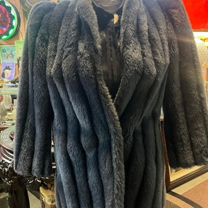 Puede incluir: Un abrigo de piel sintética gris oscuro con un cuello ancho y mangas largas. El abrigo tiene un diseño vertical texturizado y se exhibe en un maniquí. El abrigo parece ser de estilo vintage o retro.