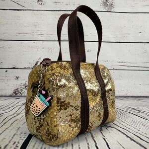 May include: A small, gold-coloured handbag with a textured, scale-like pattern. The bag has brown handles and a matching strap. A charm of a bubble tea drink hangs from the zipper. The bag is on a light-coloured wooden surface.