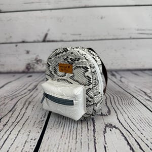 May include: A small backpack with a snakeskin pattern in black and white. The front pocket is white with a gray zipper. The bag has brown straps and a small brown leather tag.