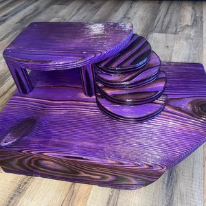 May include: Purple wooden tiered stand with a flat top and a curved base. The stand is made of multiple layers of wood, each stained a different shade of purple.