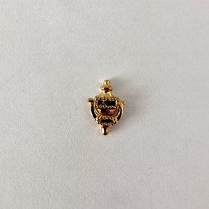 May include: A small, gold-coloured door knocker charm with an intricate design. The charm has a classic door knocker shape with a ring and a decorative top. The charm is likely made of metal and is set against a white background.