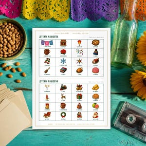 May include: A white Loteria Navide&ntilde;a game board featuring two grids of colourful Christmas-themed illustrations and Spanish text labels. The board is surrounded by a bowl of beans, a stack of blank cards, a glass bottle, sunflowers, and a cassette tape.