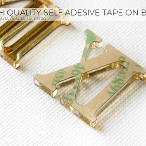 May include: Close-up of gold-colored metal letter cutouts with adhesive tape on the back. The letters are on a white textured surface. Text at the top reads "HIGH QUALITY SELF ADHESIVE TAPE ON BACK". The bottom right corner has the text "Glow Me MADE IN ITALY".