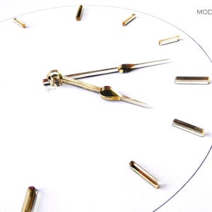 May include: A minimalist white clock face with gold hour markers and hands. The clock's hands indicate approximately 2:10. The text "MOD. 12LINES" is in the upper right corner. The brand name and "MADE IN ITALY" are in the lower right corner.