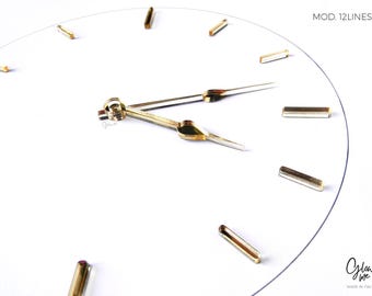 May include: A minimalist white clock face with gold-colored hour markers and hands. The clock's hands point to approximately 2:10. The text "MOD. 12LINES" is in the upper right corner. The brand name and "MADE IN ITALY" are in the lower right corner.