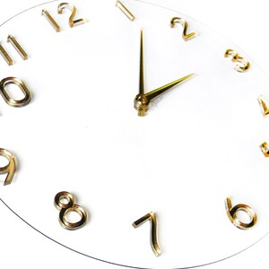 May include: A modern, minimalist wall clock with a clear face and gold-colored numerals and hands. The clock's hands point to approximately 2:10. The text "MOD. G" is in the upper left corner. The clock is a decorative home decor item.