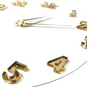 Könnte beinhalten: Eine moderne Wanduhr mit goldfarbenen Ziffern und Zeigern auf weißem Hintergrund. Das Zifferblatt zeigt die Zahlen 1 bis 5, wobei die Zeiger auf etwa 14:30 Uhr zeigen. Die Uhr ist mit "MOD. F" gekennzeichnet.