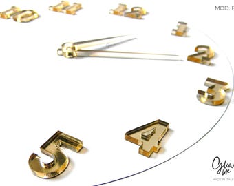 May include: A modern wall clock with gold-colored numerals and hands against a white background. The clock face features the numbers 1 through 5, with the hands pointing to approximately 2:30. The clock is labeled "MOD. F".