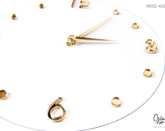 May include: A modern, minimalist clock with a clear face and gold-colored hands and hour markers. The clock face is white, and the hour markers are small, round, and gold-toned. The hands are also gold-colored. The text "MOD. 4GD" is visible.