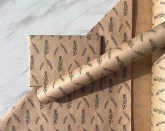 May include: A roll and a flat sheet of light brown kraft wrapping paper featuring a repeating dark green pattern of small pine needle sprigs. The paper is displayed in a rolled format and a rectangular folded sheet, both showing the same botanical print.