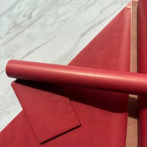 Op de afbeelding: Een bovenaanzicht van rood cadeaupapier. Een rol rood cadeaupapier op de voorgrond, met een vel rood papier en meer rollen op de achtergrond. Het papier heeft een subtiel textuurpatroon en een lichte glans.