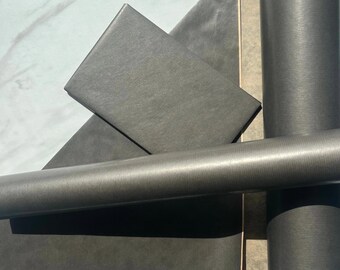May include: A dark grey rectangular box and a matching cylindrical tube, both featuring a fine, linear textured finish. The items are arranged in a geometric composition, showcasing a consistent matte dark grey surface across all visible parts.