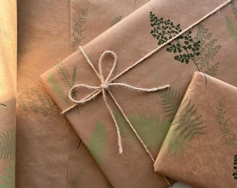 May include: Brown kraft wrapping paper featuring a repeating pattern of dark green and light green fern leaf illustrations. The paper is folded around a rectangular object and secured with a simple bow made of thin, natural jute twine.