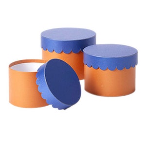 May include: Three round gift boxes with scalloped edges. The boxes are made of brown cardboard with blue lids.