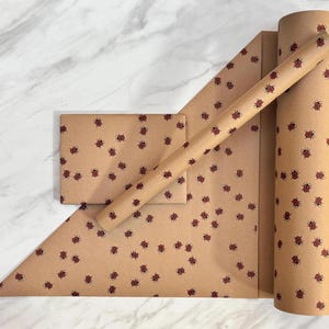 Puede incluir: Un rollo de papel de regalo de color marrón claro y una tarjeta rectangular a juego, ambos con un patrón repetitivo de pequeñas mariquitas rojas y negras dispersas por la superficie en una orientación constante y uniforme.