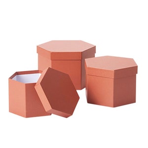 May include: Three hexagonal gift boxes in a terracotta color. The boxes are nested inside each other, with the largest box on the bottom and the smallest box on top. The boxes are made of cardboard and have a smooth finish.