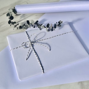 May include: A wrapped gift in white paper, tied with silver and white twine. A rolled sheet of white paper and a sprig of eucalyptus are also visible. The items are arranged on a white surface.