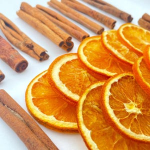 May include: A collection of dried orange slices and whole cinnamon sticks arranged on a white surface. The orange slices are translucent with a vibrant orange hue, while the cinnamon sticks are brown, woody, and cylindrical in shape.