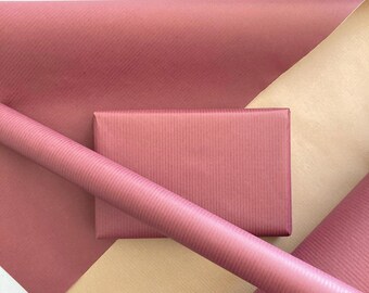 May include: A rectangular gift box wrapped in dusty rose paper with a fine vertical ribbed texture, positioned over a matching roll of wrapping paper and a sheet of light beige paper, creating a layered composition of paper goods.