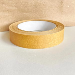 Peut inclure: Un rouleau de ruban adhésif en papier de couleur beige avec un cœur blanc. Le ruban est déroulé, révélant le côté adhésif. L'arrière-plan est une surface claire et un sac en papier brun.