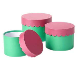 Può includere: Tre scatole regalo rotonde con bordi a festoni. Le scatole sono verdi con coperchi rosa.