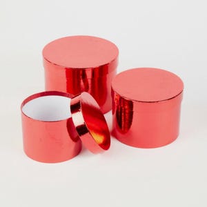May include: Three red, cylindrical gift boxes with shiny, metallic surfaces. One box is open, revealing a white interior. The lids are also red and metallic. The boxes are of varying sizes, with one lid slightly ajar.
