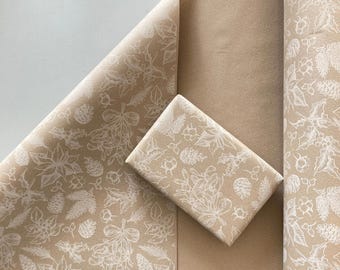 May include: A roll of light brown kraft wrapping paper featuring a repeating white line art pattern of mistletoe, pinecones, and holly leaves. A rectangular gift box wrapped in the same paper is placed on top of the unrolled paper sheet.
