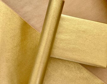 May include: A roll of metallic gold wrapping paper with a fine, horizontal textured finish. The paper is partially unrolled, revealing a smooth, reflective surface that catches the light, creating subtle variations in the golden tone.