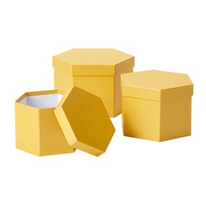 Può includere: Tre scatole regalo esagonali gialle con coperchi. Le scatole sono in cartone e hanno una finitura liscia.