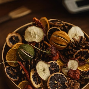 Könnte beinhalten: Eine Holzschale gefüllt mit getrockneten Fruchtscheiben, Tannenzapfen und roten Chilischoten. Das Obst umfasst Orangen, Äpfel und eine grüne, runde Frucht. Die Anordnung ist eine Mischung aus Texturen und Farben, die eine natürliche, rustikale Ästhetik erzeugt.