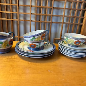 May include: A vintage tea set with floral designs. The set includes a creamer, teacups, saucers, and a stack of plates. The cups and creamer have blue and purple accents with orange and yellow flowers. The plates and saucers have a blue border.