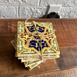 May include: Set of four yellow ceramic coasters with a blue, green, and red geometric pattern. The coasters are tied together with twine.