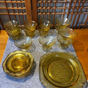 May include: A collection of vintage amber-colored glassware, including plates, bowls, and stemmed dessert dishes. The glassware features a decorative embossed pattern. The set is arranged on a gray surface, with a wooden lattice background.