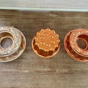 May include: Three copper-colored vintage jello molds. Two are ring-shaped with decorative patterns, and one is a round, fluted mold. The molds are arranged on a wooden surface, showcasing their intricate designs and warm tones.