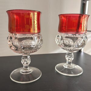 May include: Two clear glass goblets with a red, textured rim. The goblets have a round, clear base and a stem with a decorative pattern.