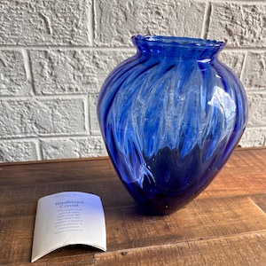 May include: A cobalt blue handblown glass vase with white swirls. The vase is sitting on a wooden surface. The text on the tag reads "Handblown Crystal. The exquisite hand-blown crystal vase is a unique collection of style and a source of lasting tradition of excellence."