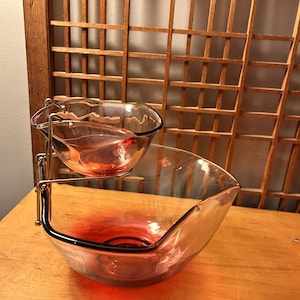 May include: A two-tiered serving bowl set. The larger, lower bowl is a clear glass with a red gradient, and the smaller bowl sits above it, held by a silver metal frame. The bowls are on a wooden surface.