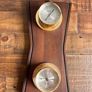 May include: A wooden wall-mounted barometer and thermometer set with gold-toned bezels. The thermometer reads in Fahrenheit and the barometer reads in inches of mercury.