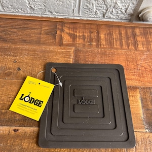 May include: A dark brown silicone pot holder with a square design and the word "Lodge" embossed in the center. A yellow tag with the "Lodge" logo and product information is attached. The pot holder is on a wooden surface.