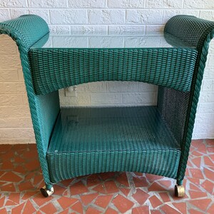 May include: A teal wicker serving cart with a glass top and a lower shelf. The cart has curved handles and sits on four small wheels. The wicker has a woven pattern.