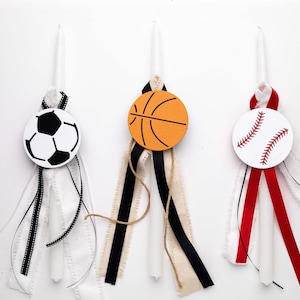 May include: Three white candles decorated with sports-themed embellishments. The candles are adorned with ribbons in black, white, red, and beige. The embellishments are a soccer ball, a basketball, and a baseball.