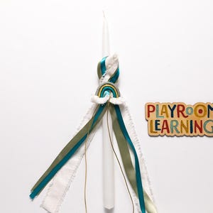 Puede incluir: Una vela blanca con una cinta de colores arcoíris atada alrededor. La cinta está atada en un nudo y tiene un pequeño amuleto de colores arcoíris adjunto. La vela está sobre un fondo blanco. El texto "PLAYROOM LEARNING" está en un letrero de madera.