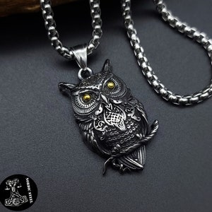 Op de afbeelding: Een zilveren ketting met een gedetailleerd zwart uilenhanger met gouden ogen. De uil zit op een tak.